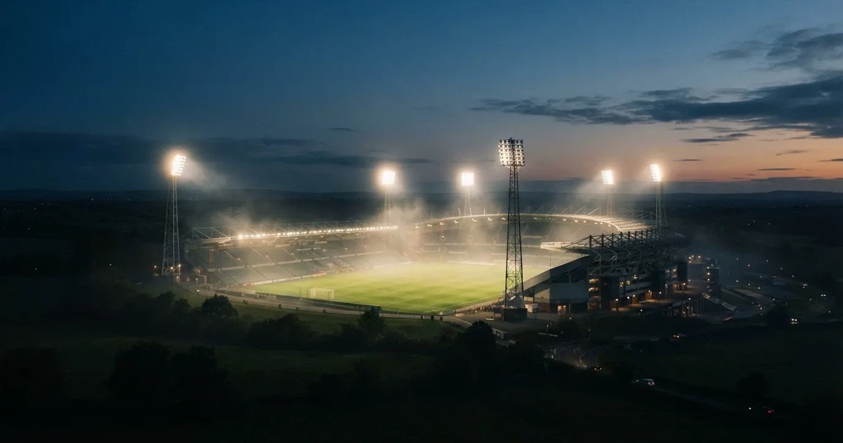 Ein Stadion unter Flutlicht aus der Ferne am Abend, hell erleuchtet, Nebel liegt über dem Rasen.
