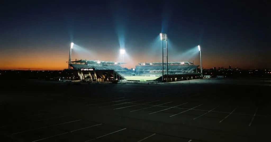 Ein Fußballstadion unter Flutlicht aus großer Entfernung gesehen, die Lichtkegel stehen im dunklen Abendhimmel über leeren Parkplätzen.